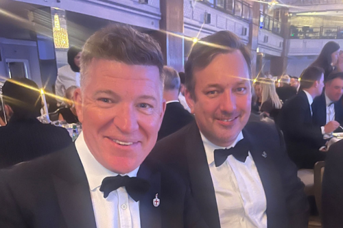 Two people in black-tie attire seated at a formal dinner event, with guests and warm decorative lighting in the background.