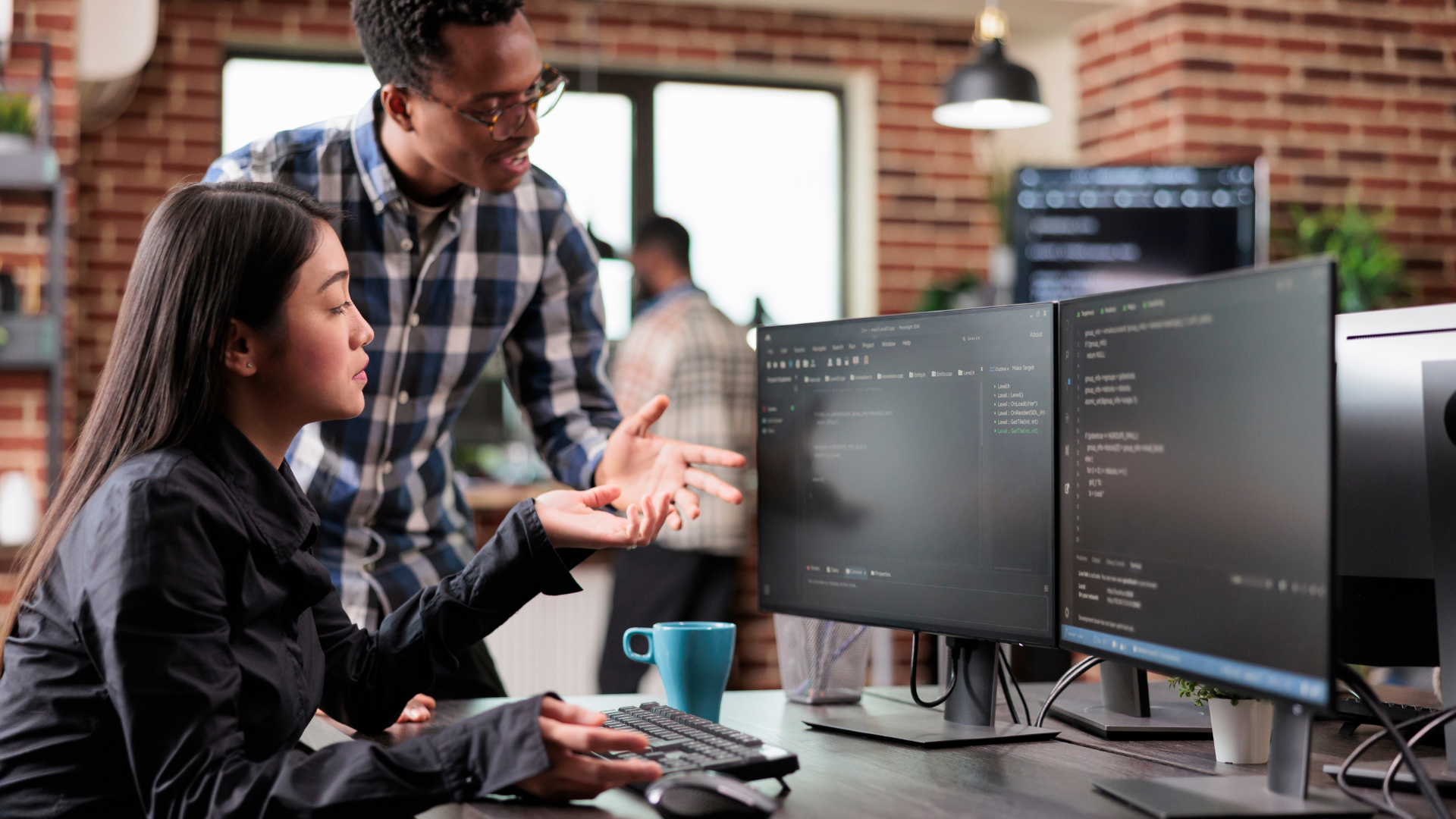 Two software developers collaborating in front of dual monitors displaying code, engaged in a discussion within a modern office setting with exposed brick walls.
