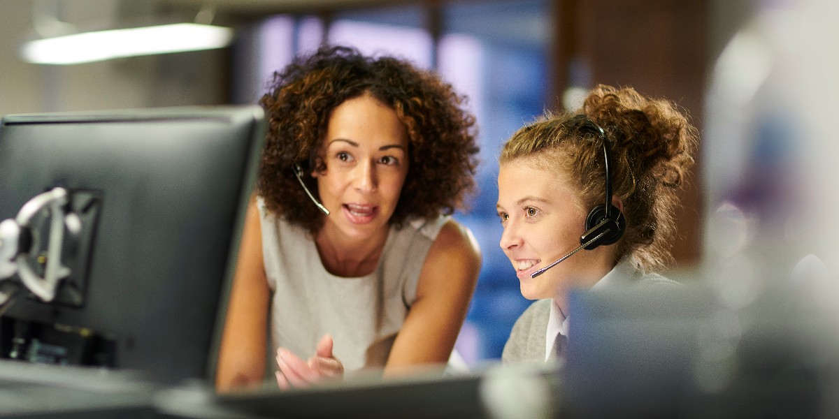 Two women wearing headsets sit together at a computer. The woman on the left, with curly brown hair, gestures toward the screen while speaking to the woman on the right. The woman on the right, with her hair in a bun, smiles while looking at the computer screen.