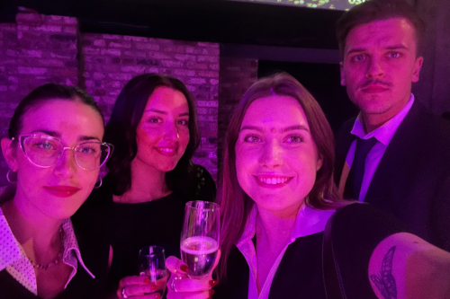Group selfie at an evening networking event, featuring four attendees holding drinks in a stylish venue with purple ambient lighting and exposed brick walls. Perfect for content related to corporate events, celebrations, networking, and social gatherings.