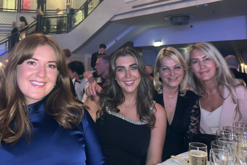 Four attendees pose together at a formal indoor event beside a table with drinks, with other guests and a balcony visible in the background.