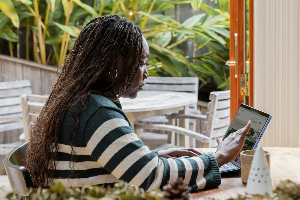A woman with long braided hair, wearing a striped jumper, sits at a wooden table using a tablet device. She is in a bright, plant-filled indoor-outdoor space with wooden patio furniture in the background.