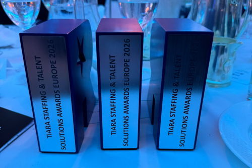 Three rectangular silver-and-black award trophies standing on a banquet table under blue lighting. The trophies display the text &ldquo;TIARA STAFFING & TALENT SOLUTIONS AWARDS EUROPE 2026.&rdquo; Wine glasses and tableware are visible in the background, suggesting an awards event.