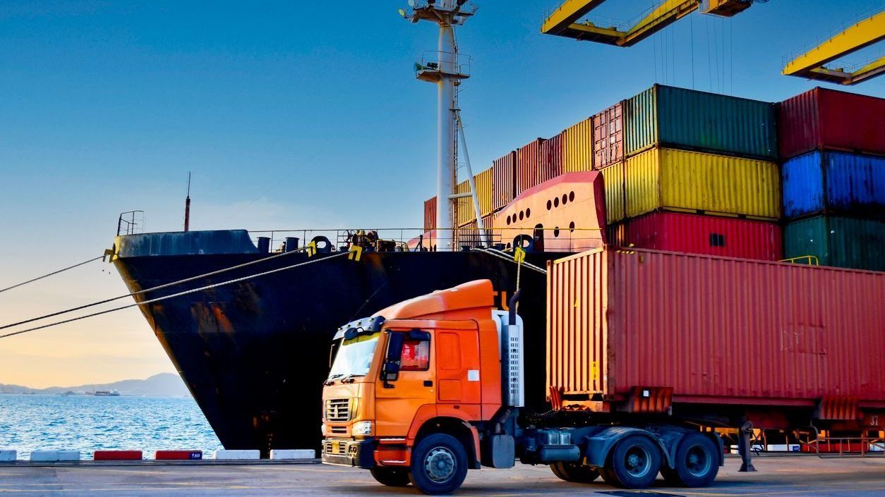 Orange semi-truck transporting a shipping container at a busy port with cargo ship and stacked containers in the background.