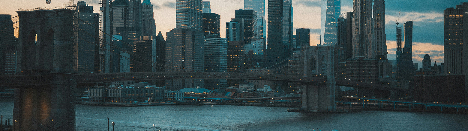 Scenic view of the Brooklyn Bridge spanning across the East River, with the illuminated Manhattan skyline in the background. The photo captures the contrast between the historic stone bridge and modern skyscrapers under a moody evening sky.