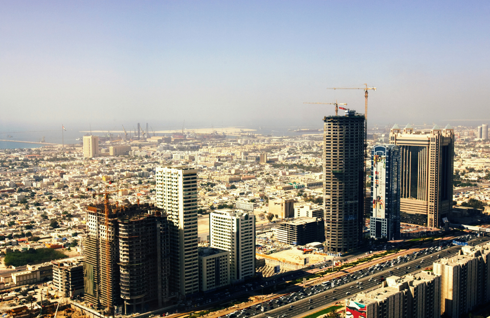 Aerial view of a dense urban skyline with high-rise buildings, several towers under construction, and a coastal port visible in the distance.