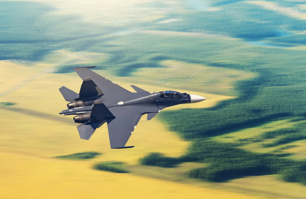 Twin-engine fighter jet flying at low altitude above patchwork fields and forested landscape, photographed from the air with motion blur in the background.