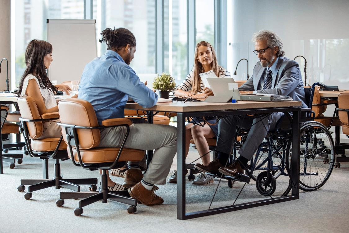 Group of professionals seated around a table in a modern office, including an individual using a wheelchair, engaged in a collaborative discussion with laptops and documents visible