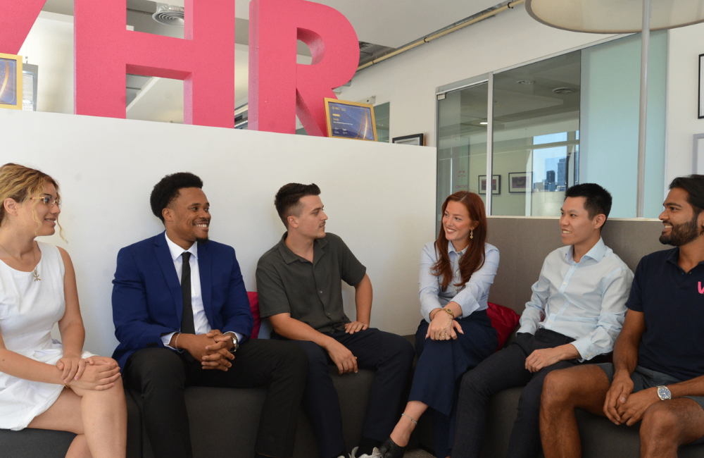 Six people seated in a modern office lounge area, engaged in conversation on a sofa. Large pink letters spelling “HR” are displayed behind a partition, with glass-walled meeting rooms and office furnishings visible in the background.