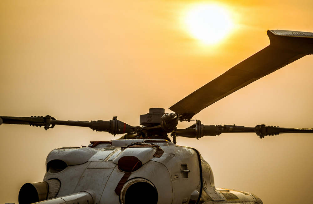 Close-up of a helicopter’s rotor mast and blades silhouetted against a golden sunset, with the bright sun positioned above the aircraft.