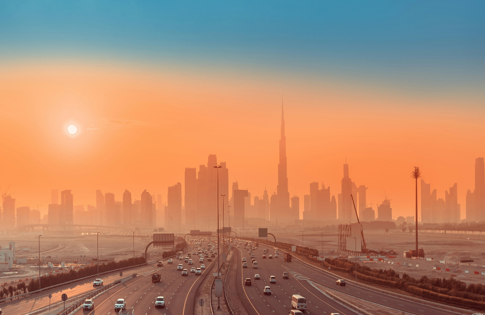 Multi-lane highway leading towards a modern city skyline at sunset, with skyscrapers silhouetted against an orange sky and heavy traffic on the road.