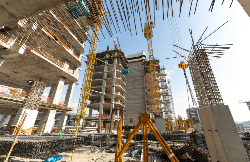 Large construction site with concrete building structures, tower cranes, exposed reinforcement steel, and ongoing high-rise development under a clear sky.