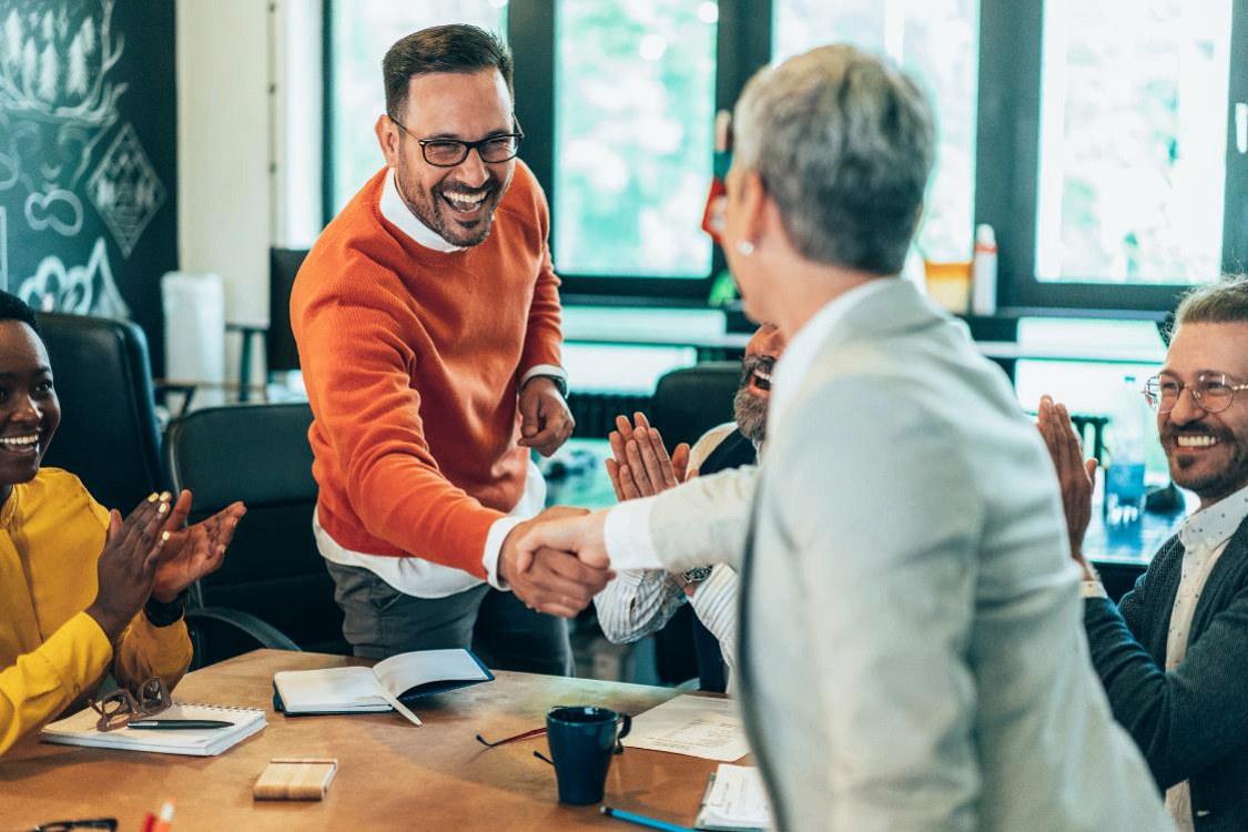 Group of professionals in a modern office shaking hands during a meeting, with notebooks and coffee cups on the table.
