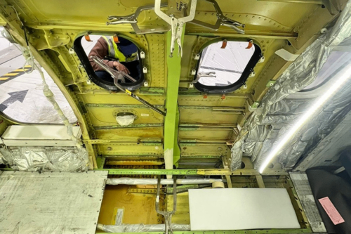 Interior view of an aircraft fuselage section with exposed structure, insulation panels, and a worker visible through an access opening performing maintenance.
