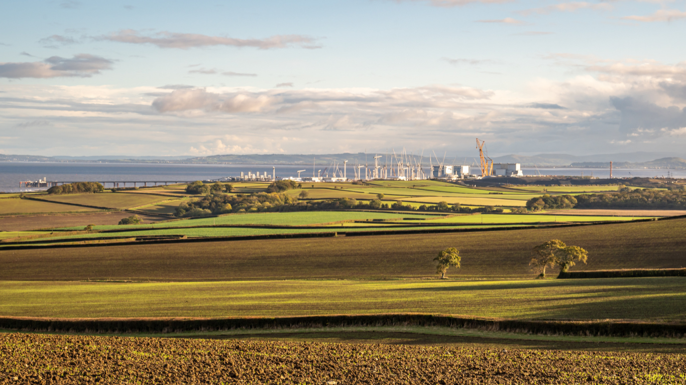 A scenic rural landscape featuring green and brown patchwork fields under a partly cloudy sky, with a large industrial construction site in the distance near the coastline. The site includes numerous cranes and tall structures, likely part of a nuclear power station development. The sea and distant hills are visible on the horizon."