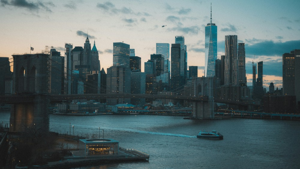 Scenic view of the Brooklyn Bridge spanning across the East River, with the illuminated Manhattan skyline in the background. The photo captures the contrast between the historic stone bridge and modern skyscrapers under a moody evening sky.