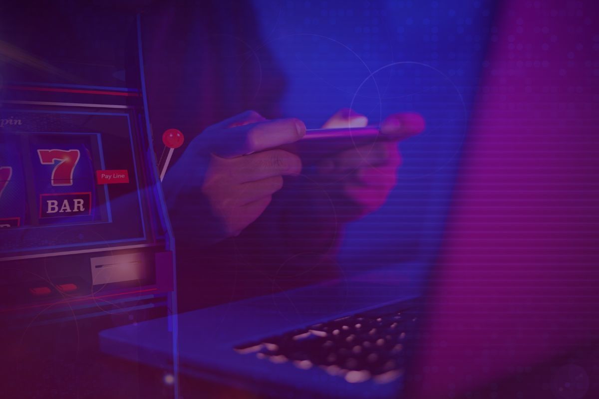 person playing games on a phone in front of a laptop, with a slots machine in the background