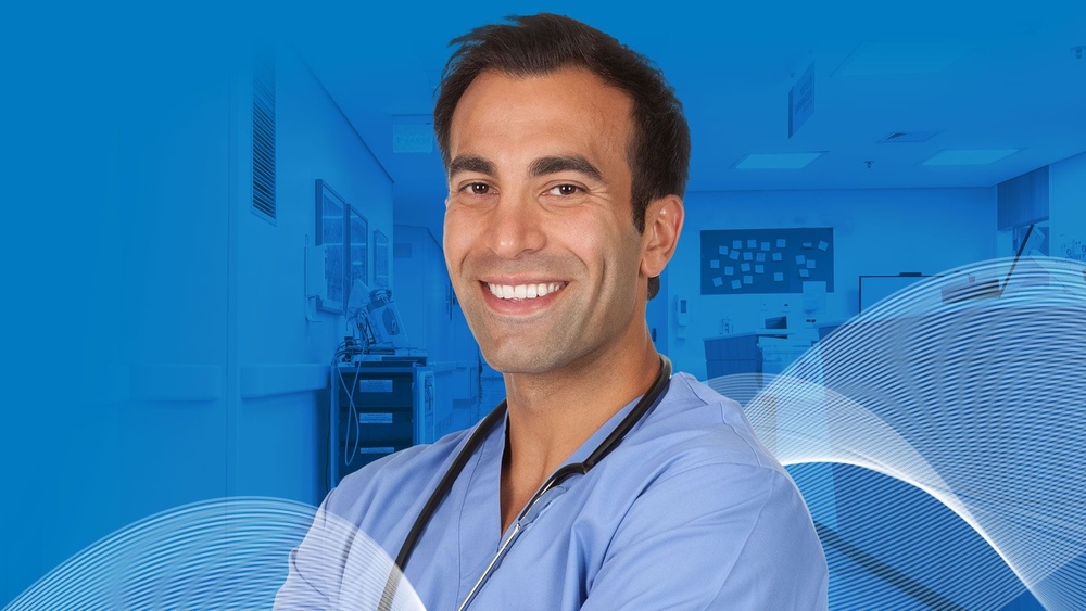 Smiling healthcare professional in blue scrubs with stethoscope, standing in a hospital corridor.