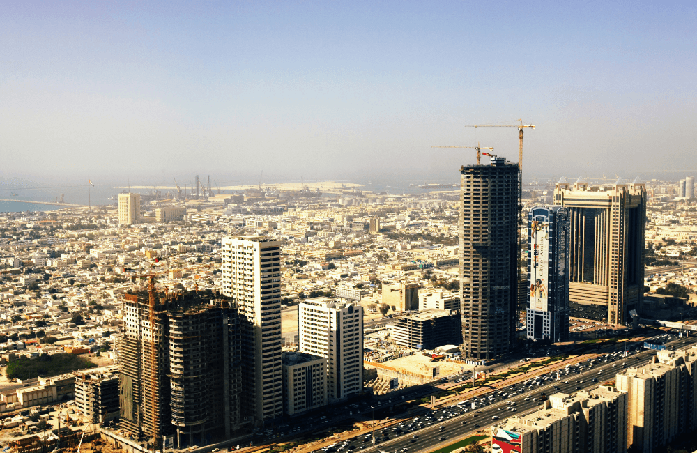 Aerial view of a dense urban skyline with high-rise buildings, several towers under construction, and a coastal port visible in the distance.