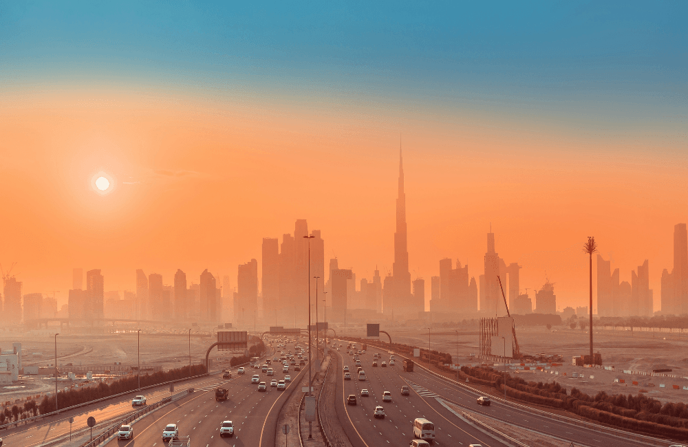 Multi-lane highway leading towards a modern city skyline at sunset, with skyscrapers silhouetted against an orange sky and heavy traffic on the road.