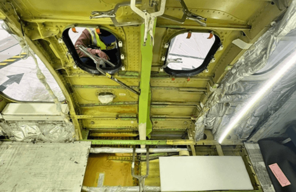 Interior view of an aircraft fuselage section with exposed structure, insulation panels, and a worker visible through an access opening performing maintenance.