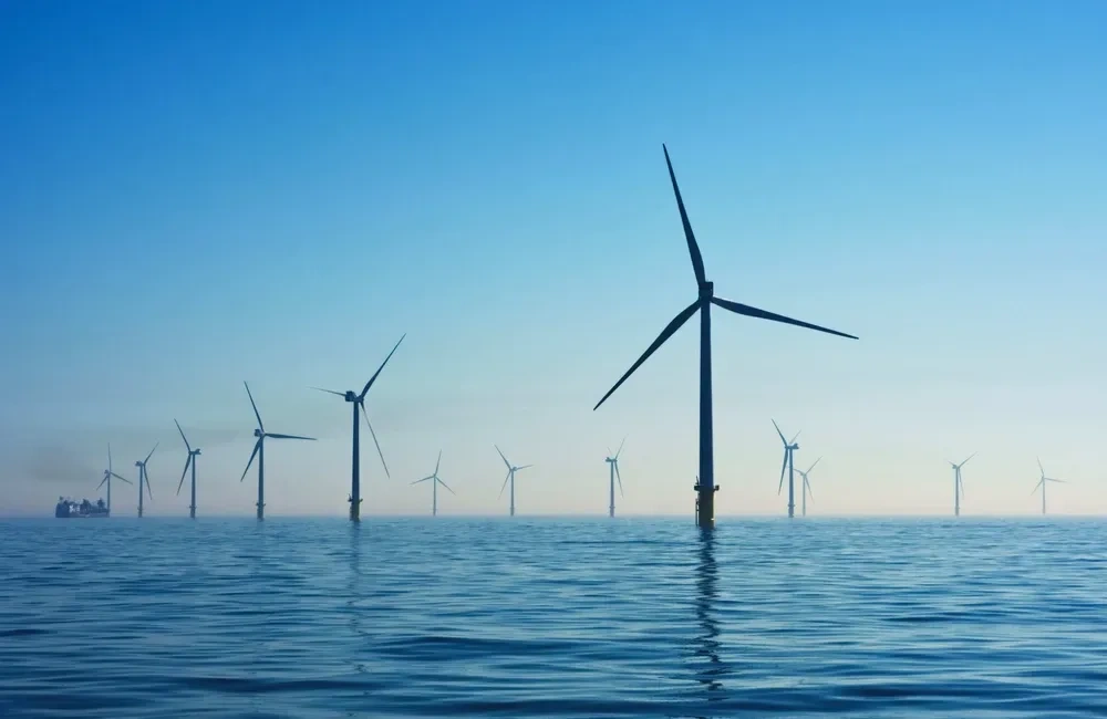 Offshore wind turbines standing in calm blue ocean waters under a clear sky, with multiple turbines extending across the horizon.