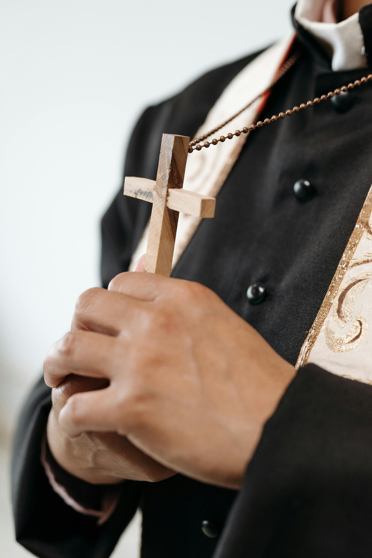 Close‑up of a person wearing dark formal clothing, holding a wooden cross necklace in both hands, with part of a light‑coloured stole or garment visible in the foreground.