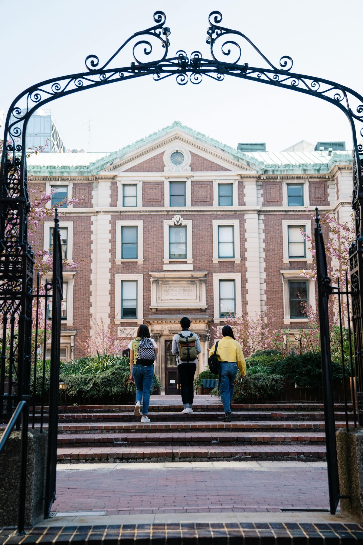 Three people with backpacks walk up steps toward a large brick academic building framed by an ornate iron gate.