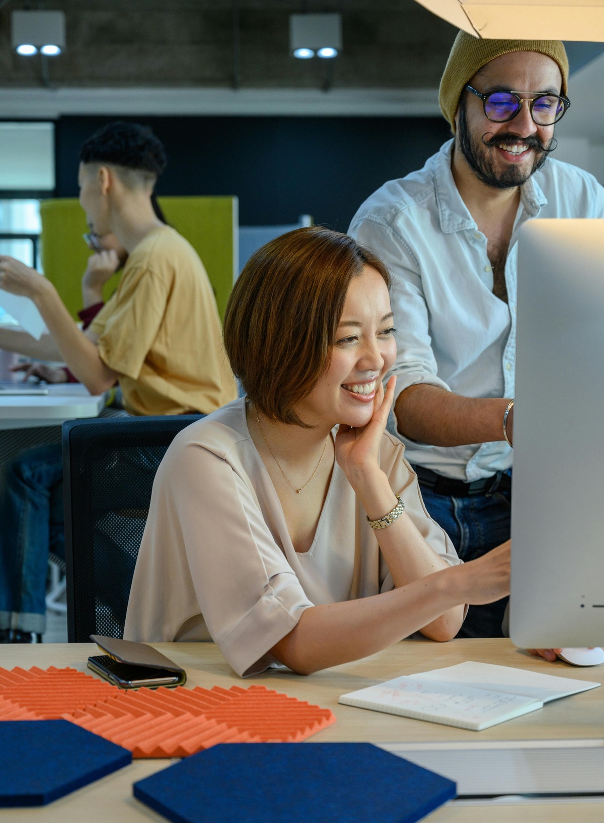 Person seated at a desk in a modern office, smiling while working at a desktop computer, with a colleague standing nearby and other people working at desks in the background.