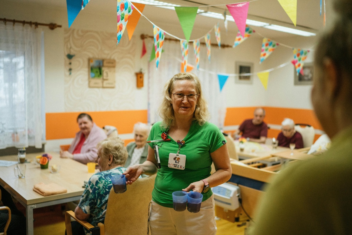 Care worker standing in a communal care home dining room, holding two cups and smiling toward someone off‑camera, with older adults seated at tables in the background and colourful bunting decorations hanging from the ceiling.