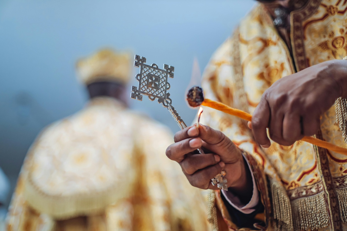 Close‑up of a person wearing ornate gold robes, holding a metal cross in one hand and a small lit candle in the other, with another similarly dressed figure softly blurred in the background.