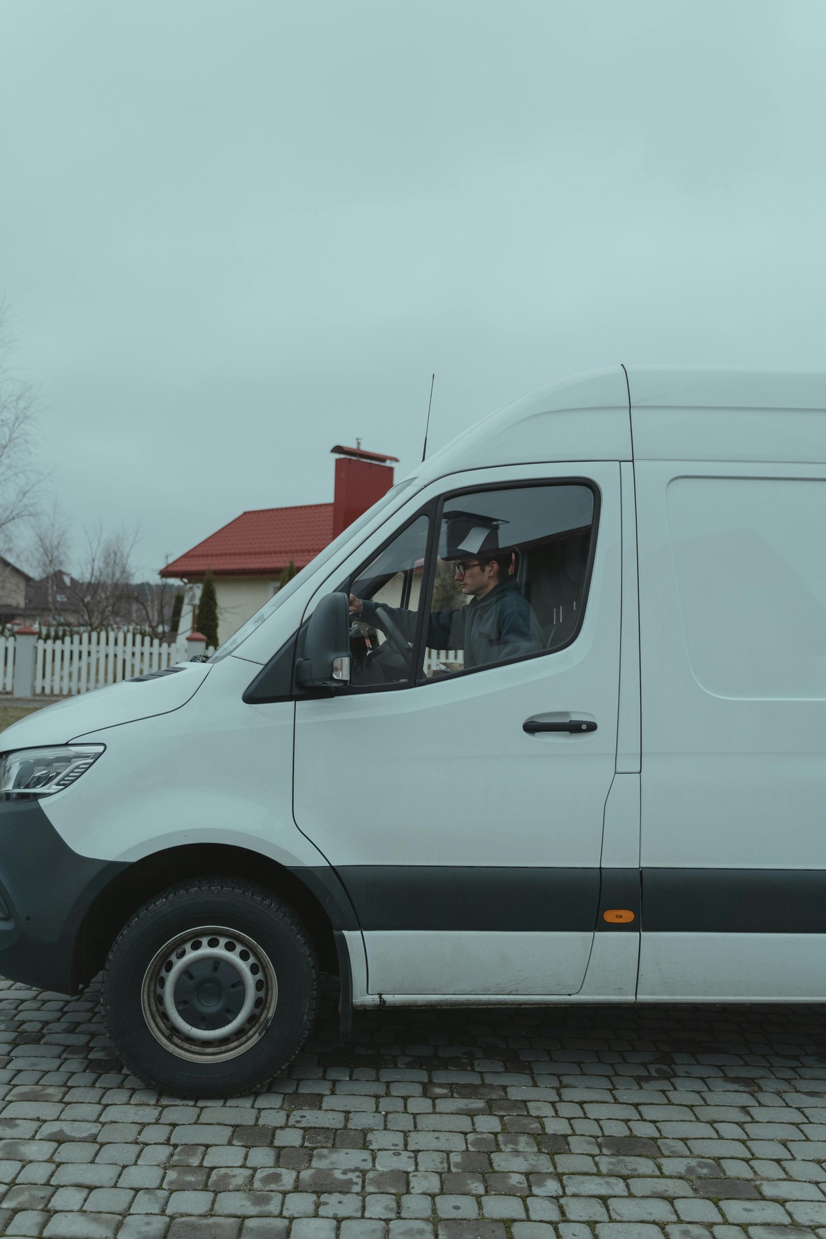 White delivery van parked on a cobblestone street, with a person seated in the driver’s seat looking forward through the open window, viewed from the side against an overcast outdoor background.