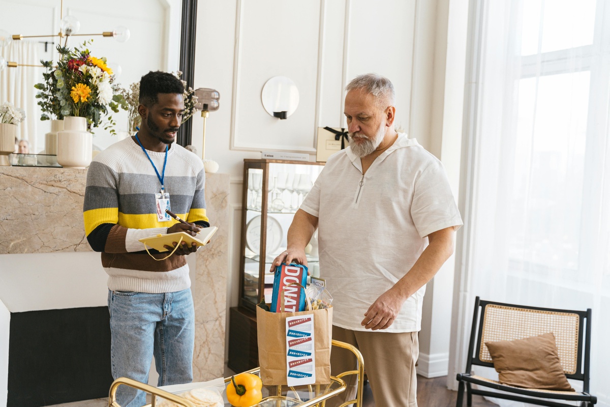 Two people standing indoors beside a table, reviewing household items, with one person holding a notepad and the other placing a boxed product into a paper shopping bag, in a bright living room with chairs, flowers, and home décor visible.