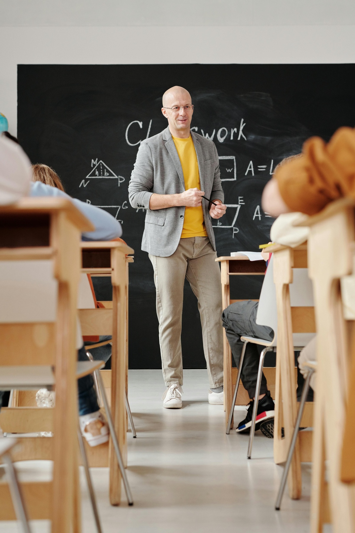 An individual stands at the front of a classroom near a chalkboard with diagrams and the word “Classwork,” while students sit at desks facing forward.