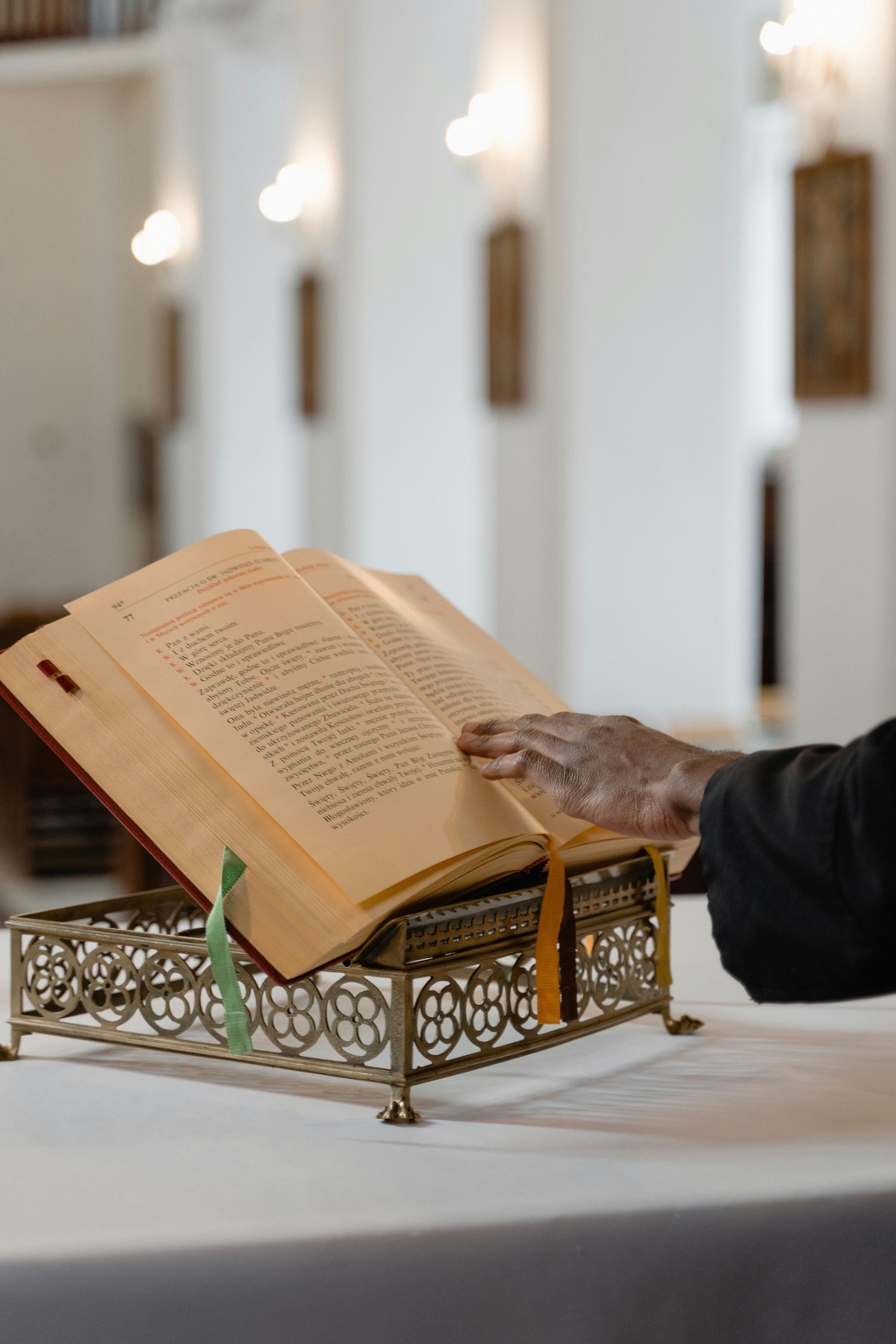 Open book resting on a decorative metal stand on a table in a bright indoor space, with a person’s hand touching the page and softly blurred architectural details visible in the background.