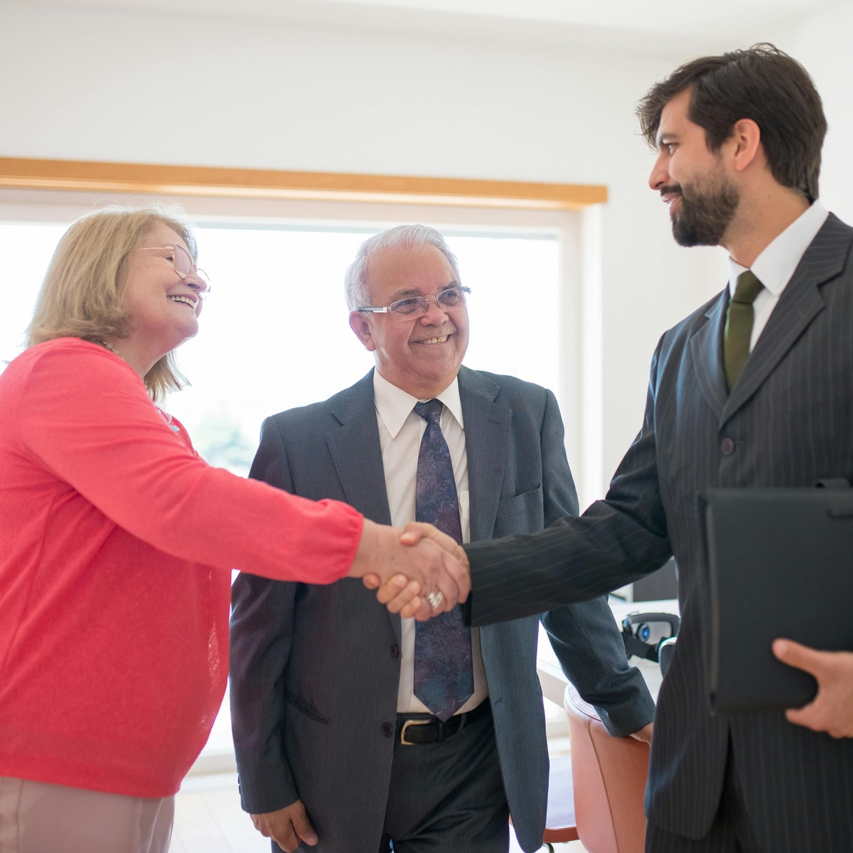 Three people stand together in an office, with two of them shaking hands while one holds a folder.