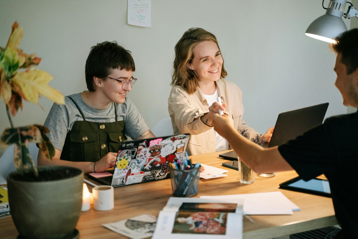 Three people seated around a desk in a creative workspace, working together with a laptop, papers, and stationery, while another person extends a hand across the table, with a desk lamp and potted plant nearby.