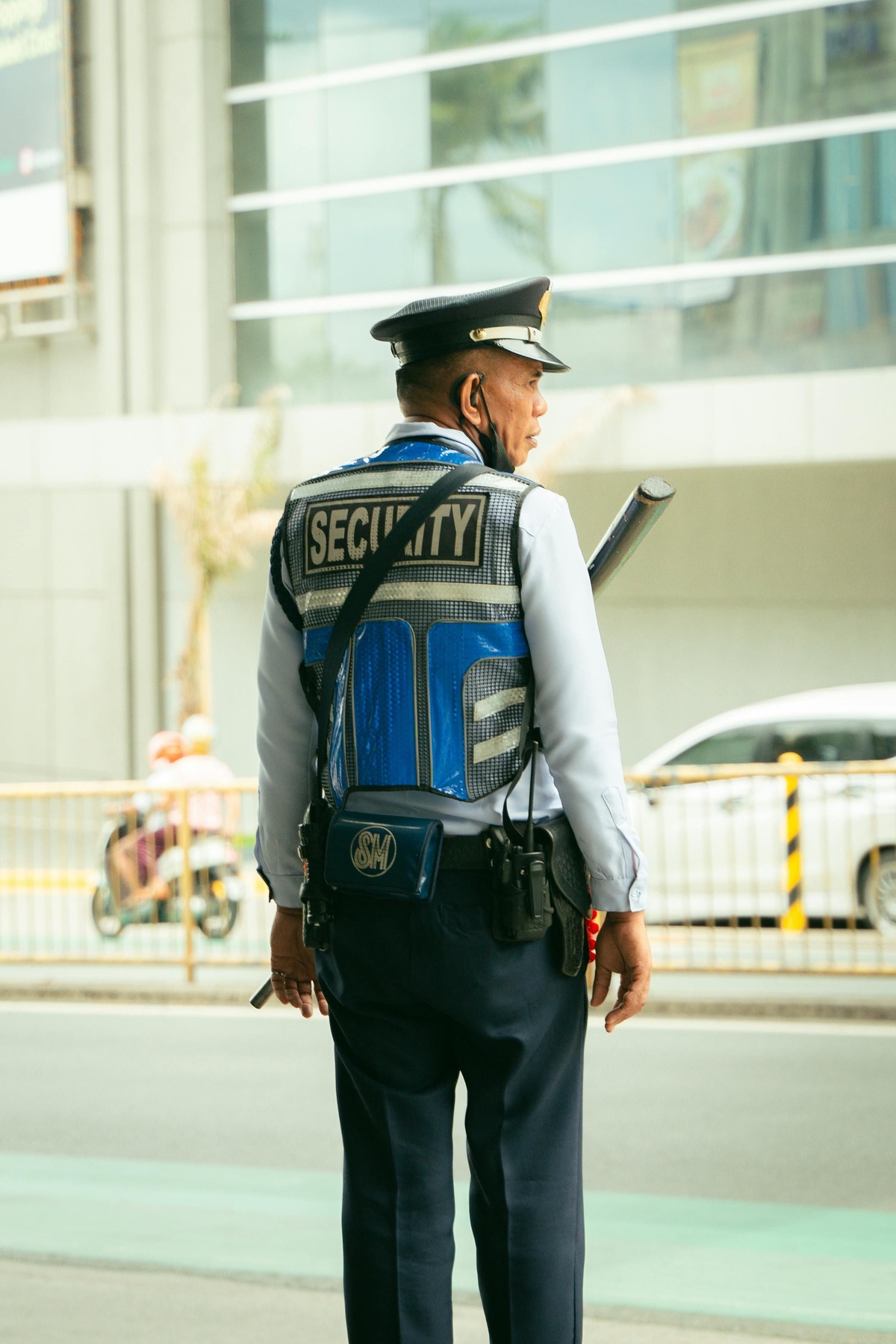 Person wearing a security uniform standing outdoors near a roadway, viewed from behind, with a vest labelled “Security,” a hat, equipment belt, and urban buildings and vehicles visible in the background.