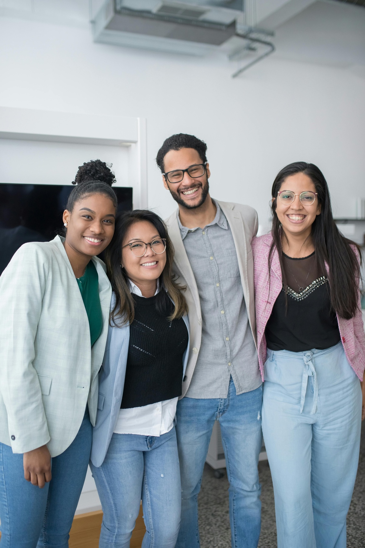 Four people standing close together in an office setting, posing side by side with arms around each other, wearing business‑casual clothing against a neutral indoor background.