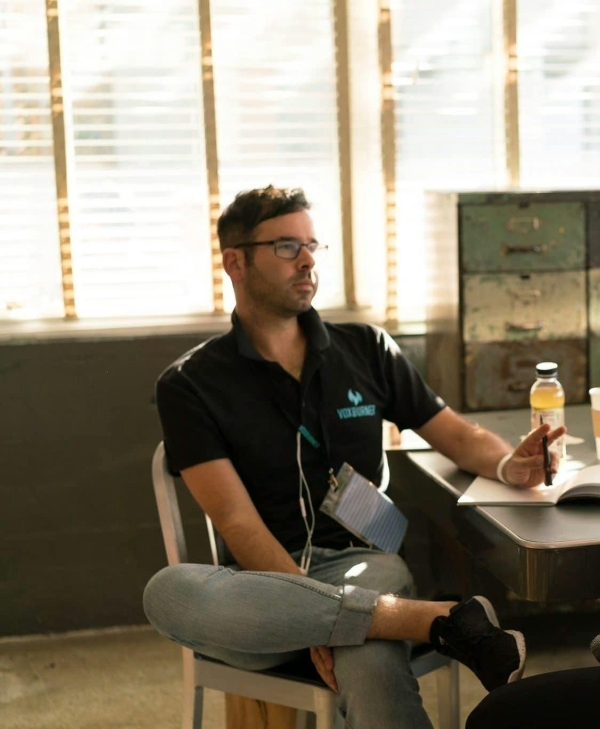 The image shows an indoor setting with a person seated on a simple chair near a desk. The person is sitting in a relaxed posture with one leg crossed over the other.