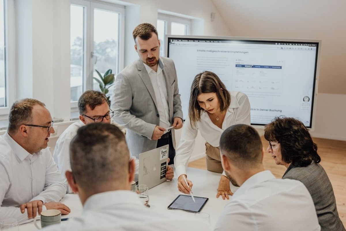 The image shows a collaborative meeting taking place in a bright, modern workspace. Several individuals are gathered closely around a table, actively engaged in what appears to be a group discussion or a working session.