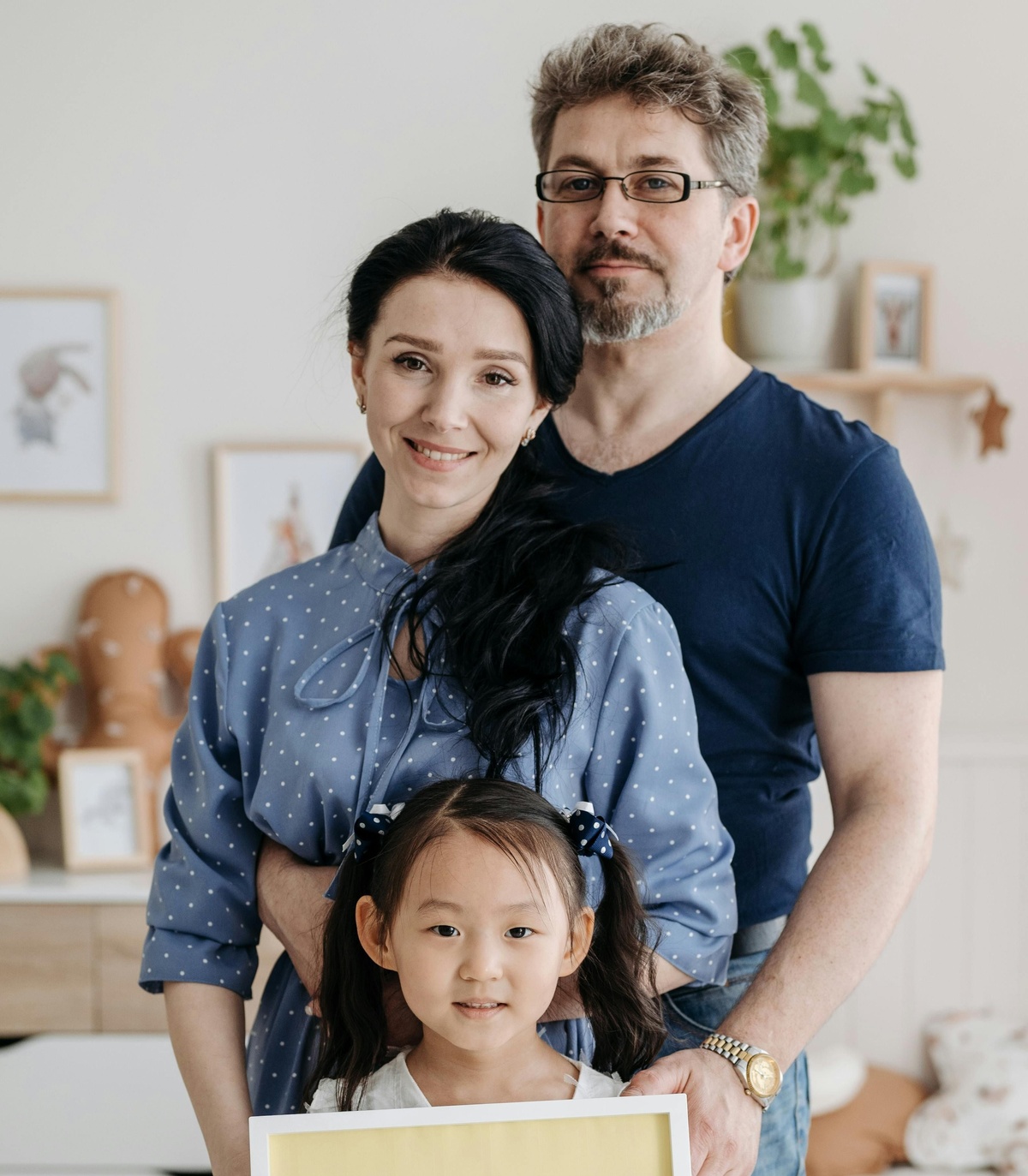Three people standing together indoors, with one smaller person holding a framed picture in the foreground and two adults standing closely behind, in a warmly lit home setting with shelves and decorations in the background.