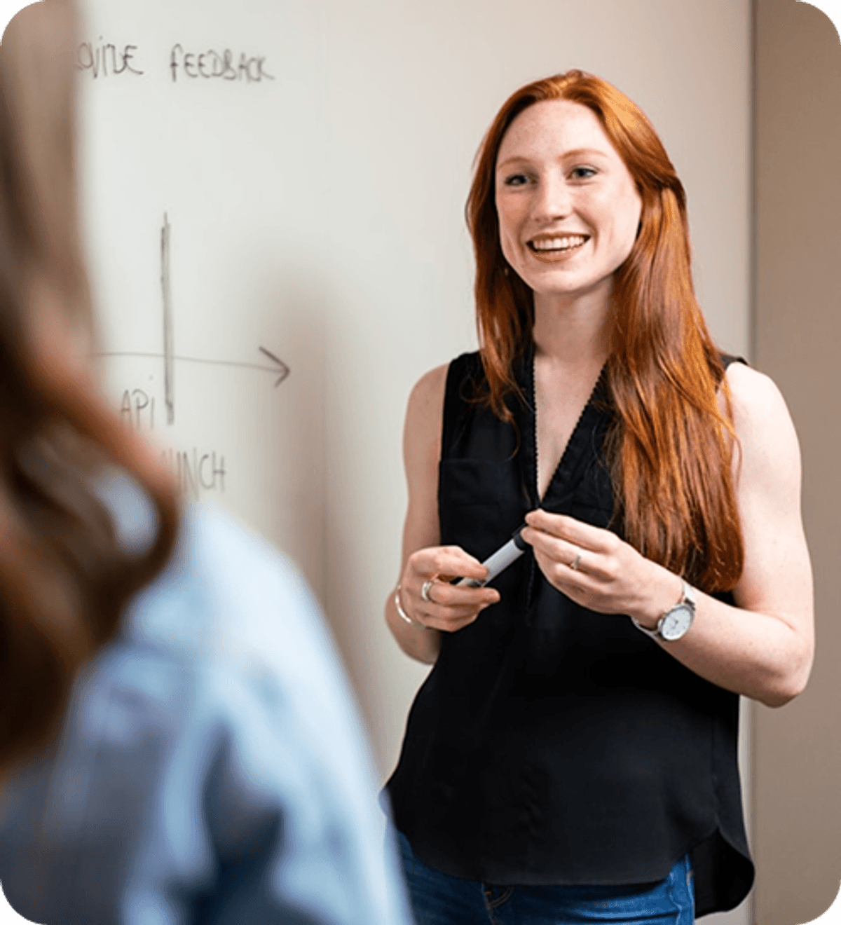 Person standing near a whiteboard, holding a marker and speaking to someone just out of frame in an indoor learning or meeting space.