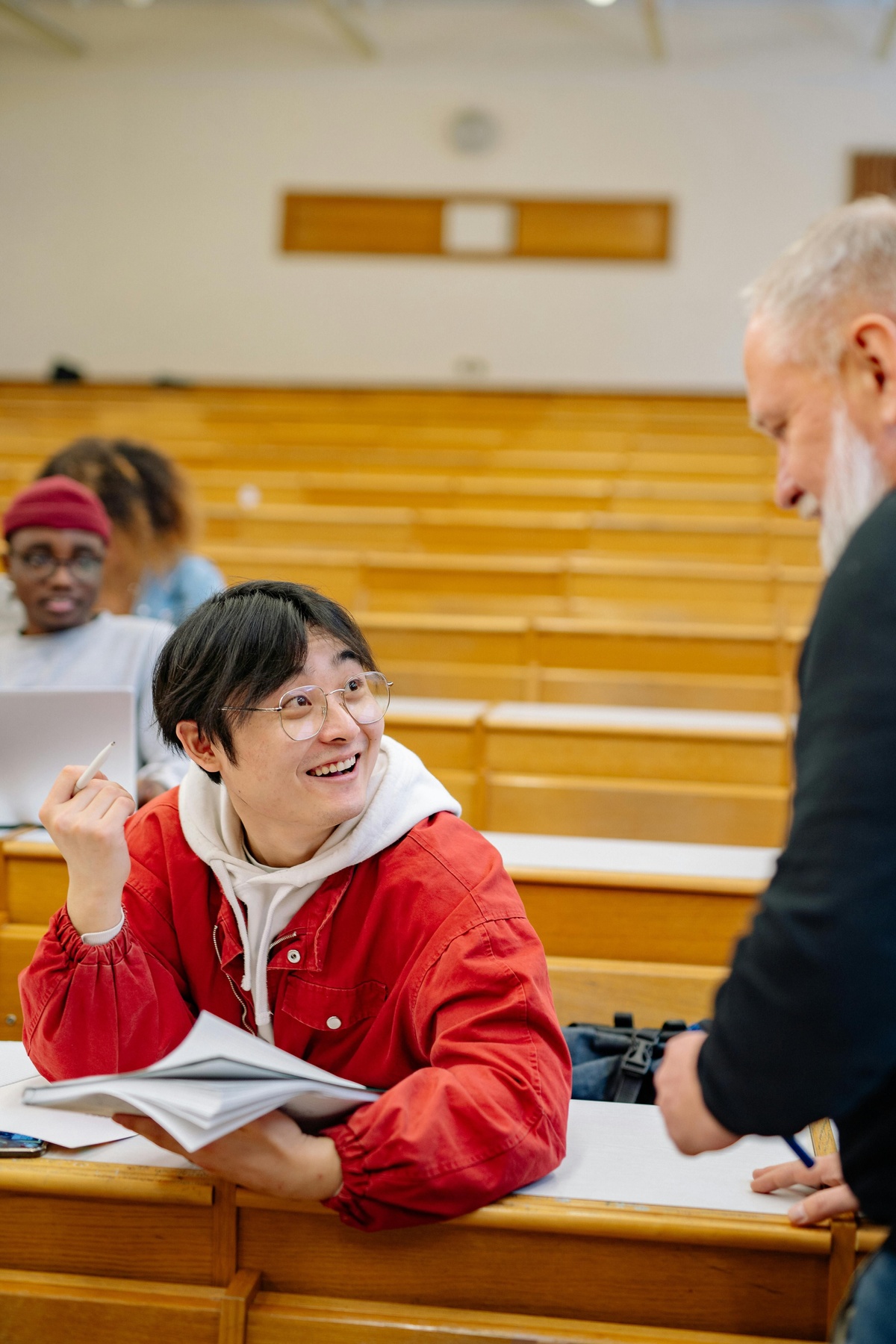 A person seated in a lecture hall holding an open notebook, while another person stands nearby during what appears to be a classroom interaction.