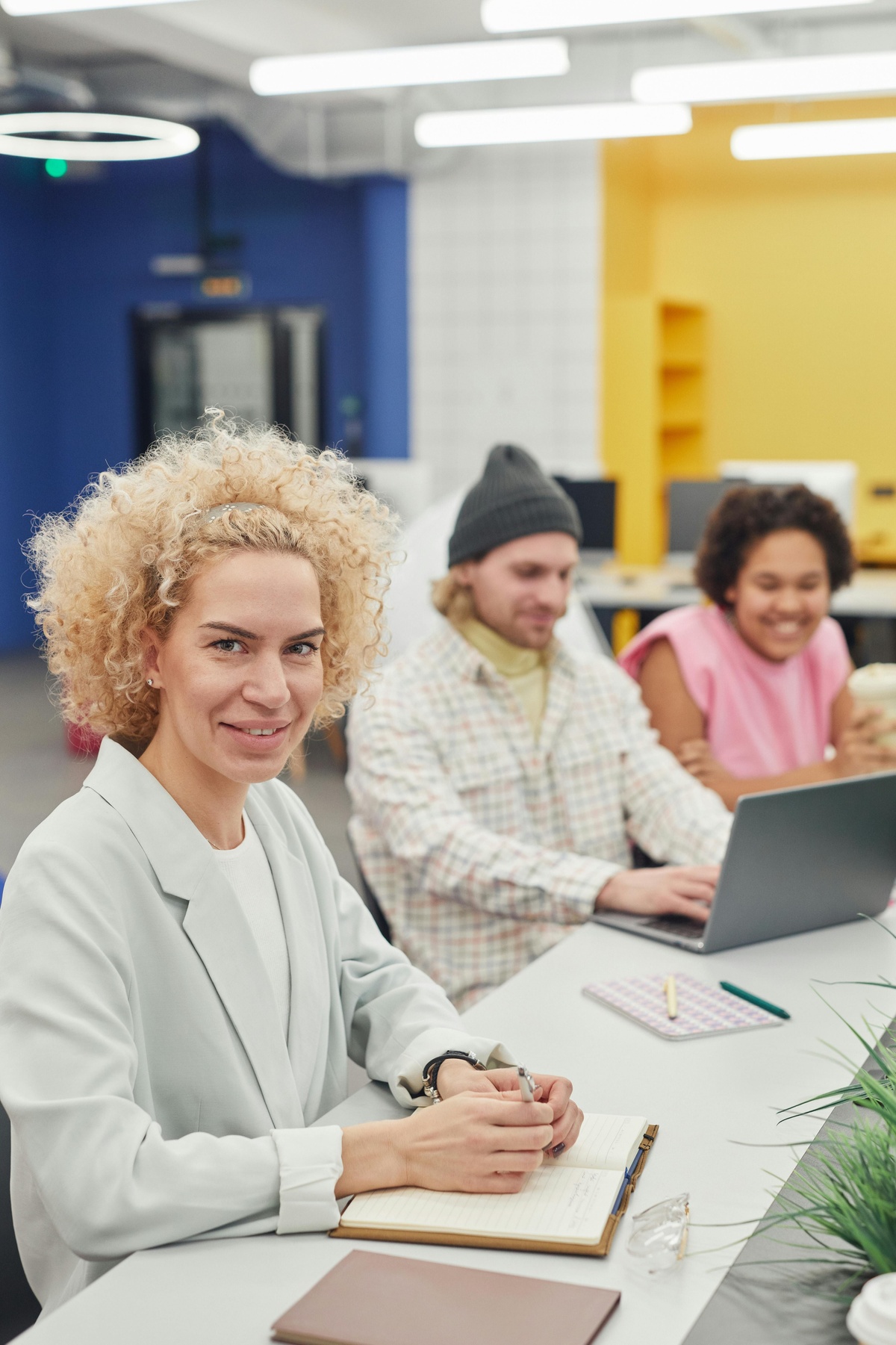 Group of people seated at a table in a modern workspace, with one person facing the camera holding a notebook while others work on a laptop in the background.