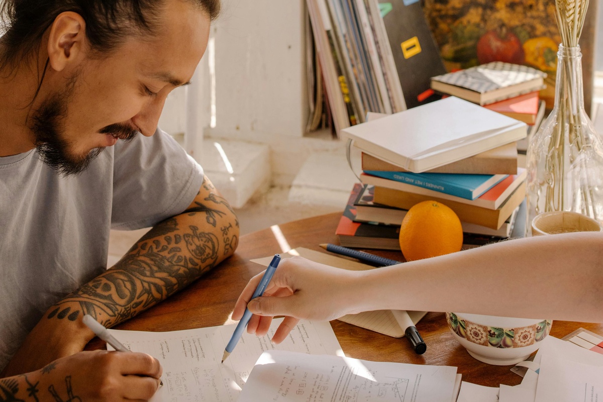 A person with tattooed arms sits at a table covered with papers while another person points at a document; books, an orange, and a patterned cup are also on the table in a warmly lit room.