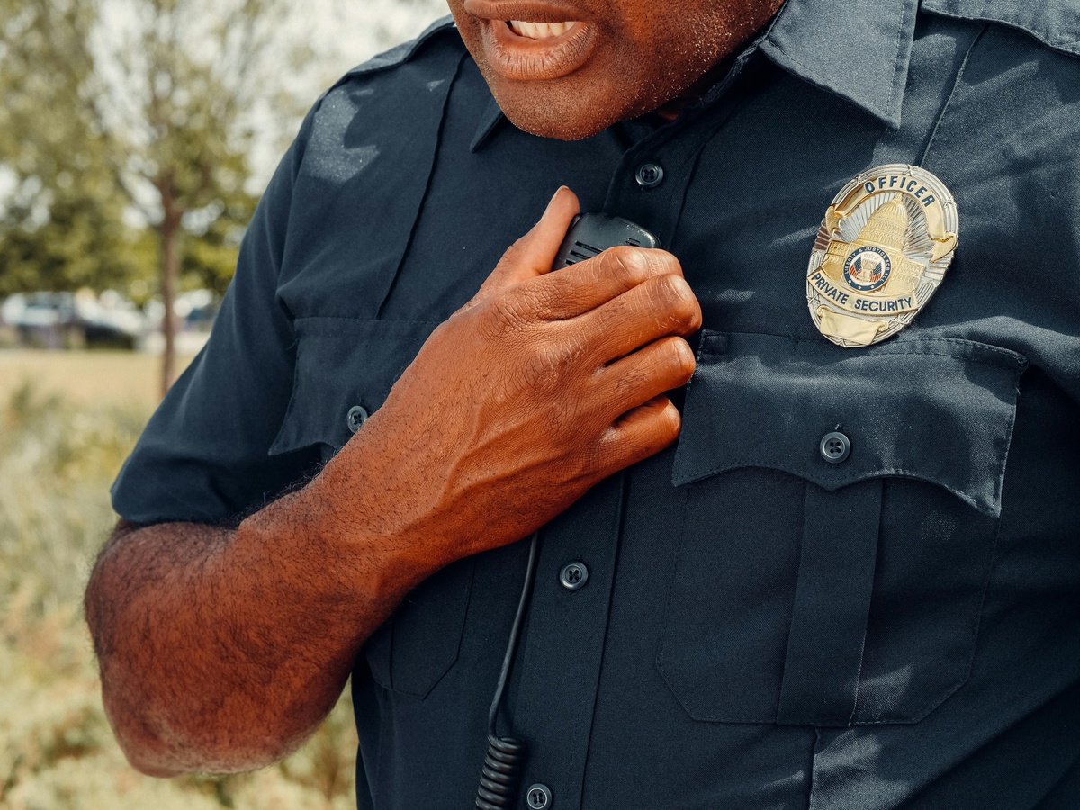 Close‑up of a police officer’s uniform, showing a hand holding a radio microphone near the chest, with a badge visible on the shirt and an outdoor background softly blurred.