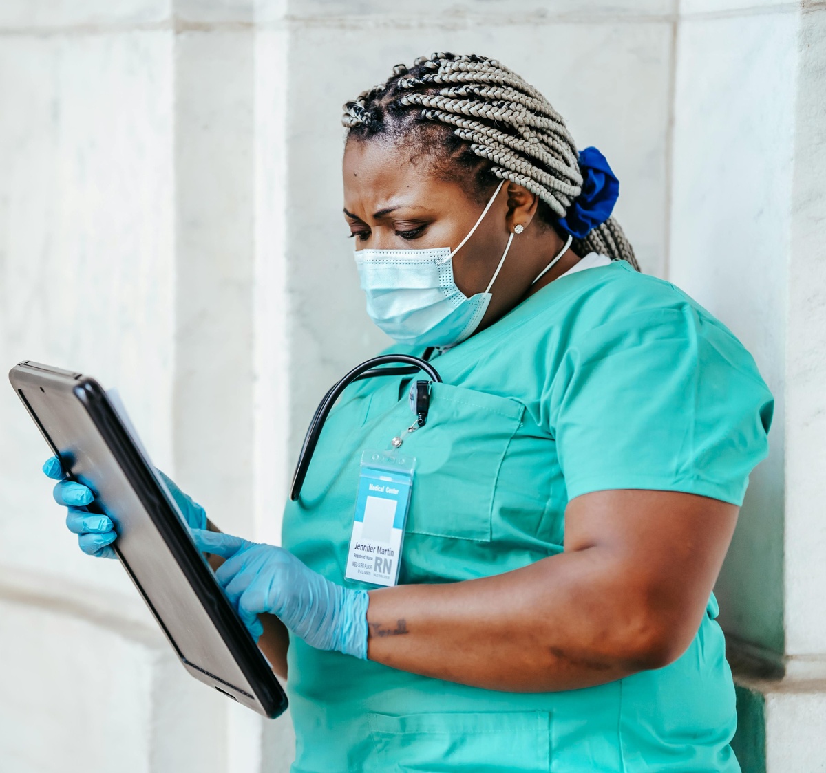 Healthcare worker wearing scrubs, gloves, and a face mask, holding and using a tablet while standing outdoors against a light‑coloured wall, with an ID badge and stethoscope visible