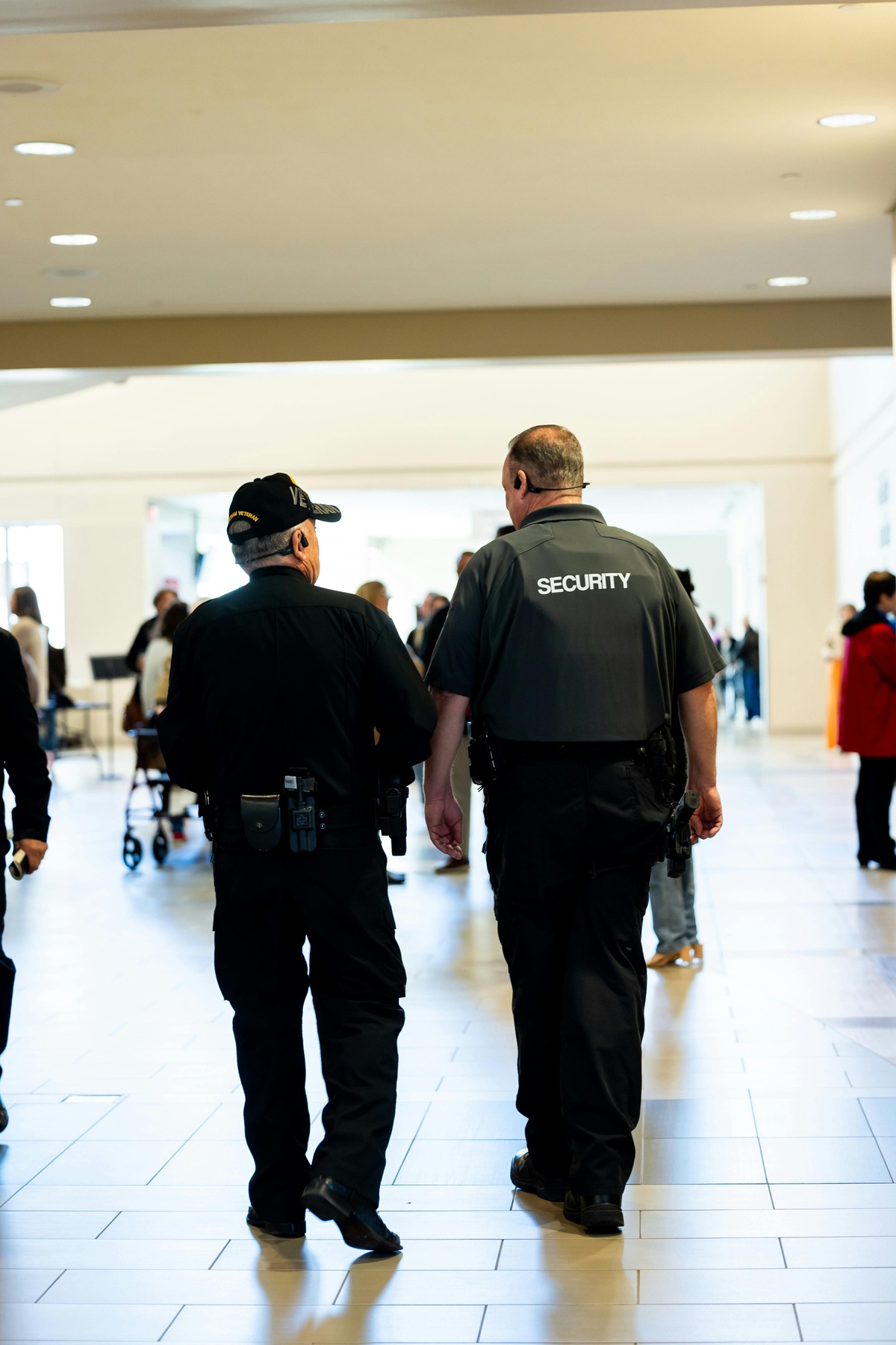 Two security personnel walking side by side through a bright indoor public space, viewed from behind, with one wearing a vest labelled “Security” and people and seating visible in the background.