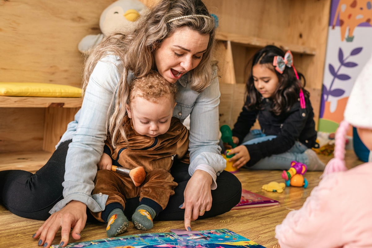 Adult and young child sitting on the floor in a playroom, looking at a colourful book together, with other children and toys visible in the background.